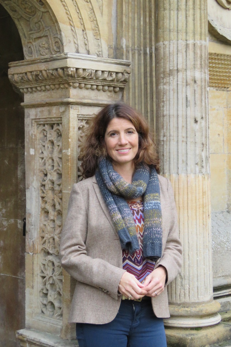 A woman wearing a brown blazer and blue and green scarf leaning against an elaborate sandstone arch
