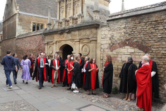 Fellows in academic gowns, mainly black and red, waiting in front of a wall