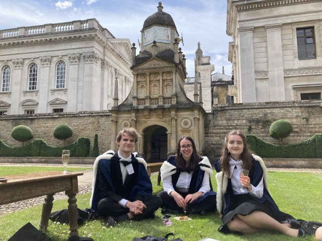 Three graduates relax on the grass in Caius Court in front of the Gate of Honour