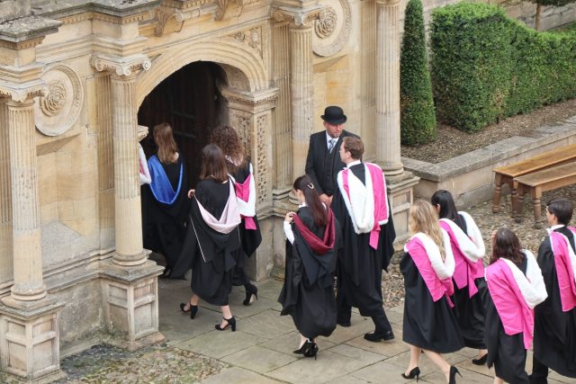 Lines of two graduates in hooded gowns pass through a narrow gateway