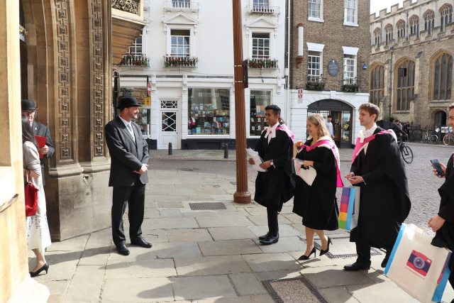 Three graduates in hooded gowns are congratulated by a Porter in a bowler hat