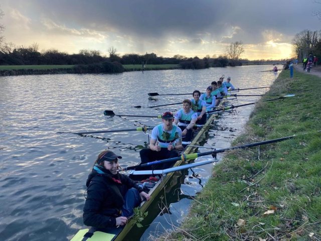 A rowing crew by a riverbank with dark clouds and sunshine overhead